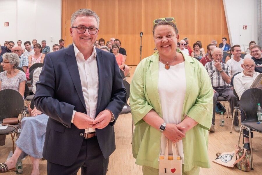 Zwei Personen stehen lächelnd vor Publikum in einem Saal.
