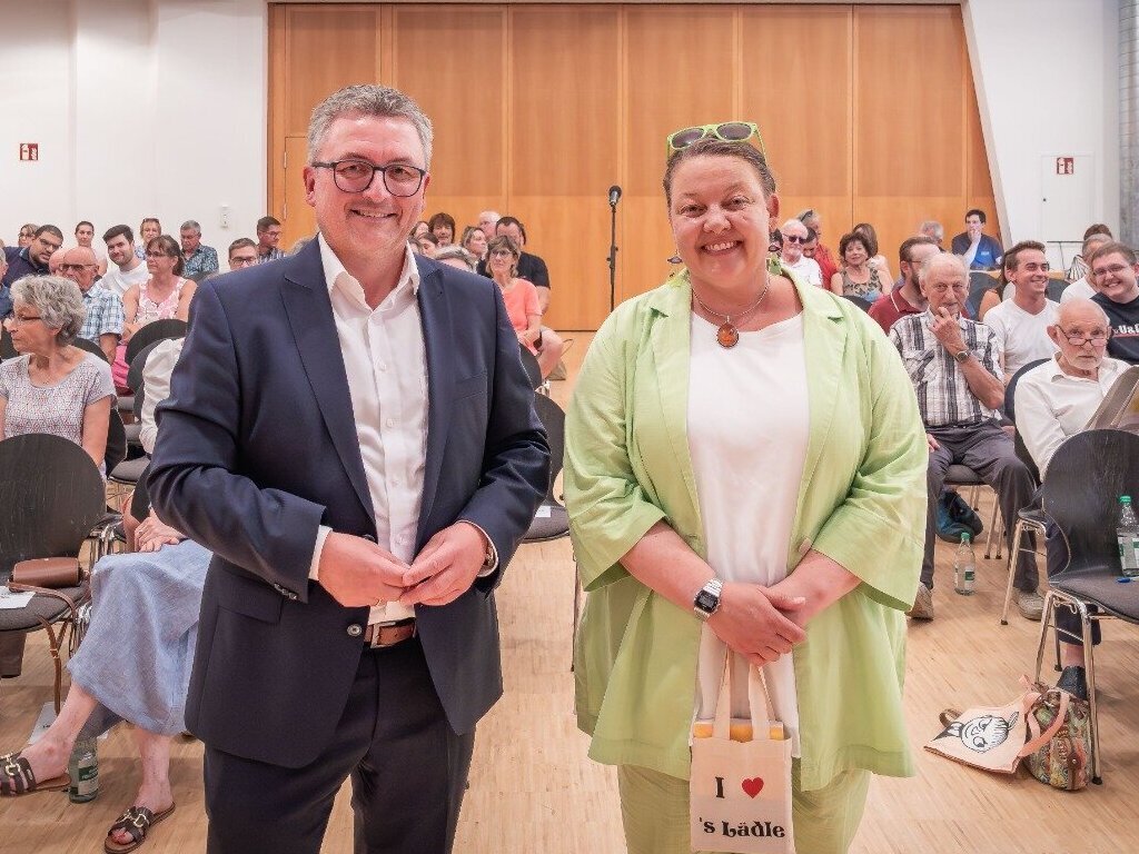 Zwei Personen stehen lächelnd vor Publikum in einem Saal.