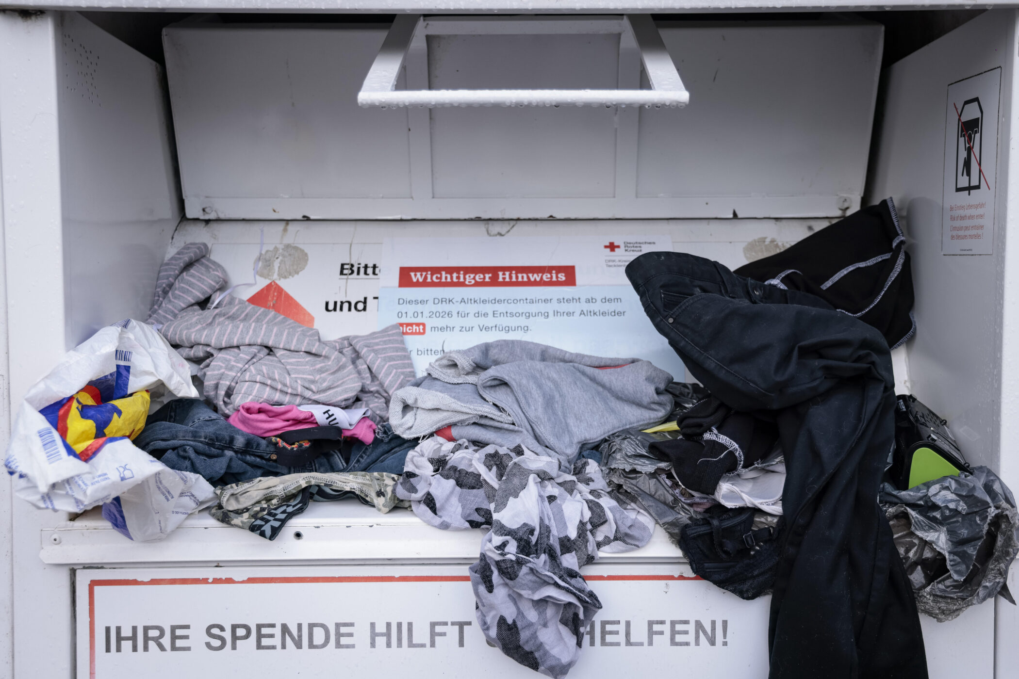 Überfüllter Altkleidercontainer mit verschiedenen Kleidungsstücken. Hinweis: Nutzung endet 2026.