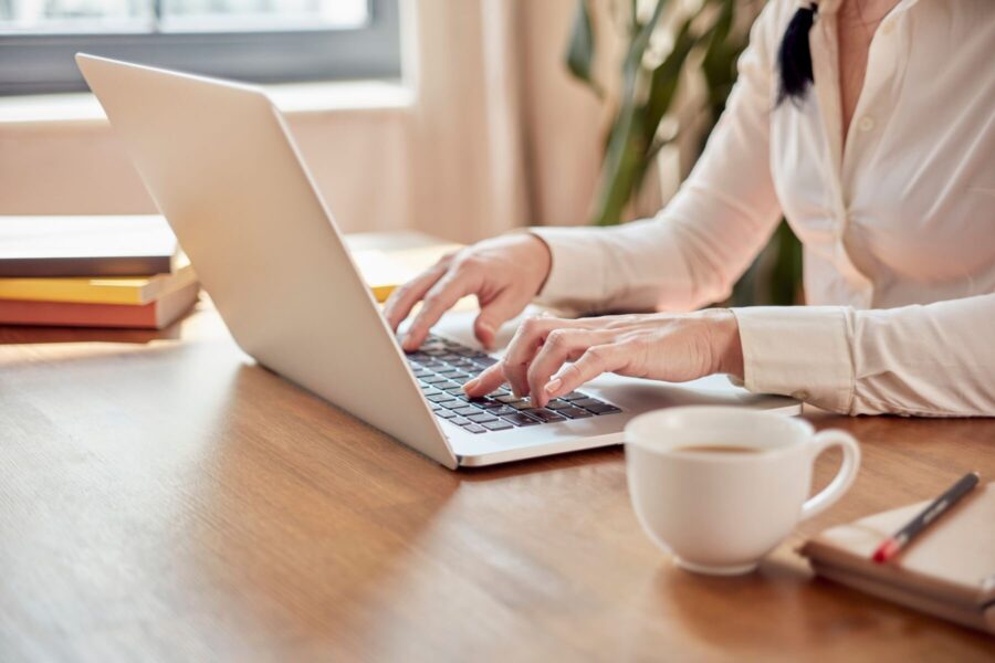 Person tippt auf Laptop, daneben eine Tasse, Bücher und Notizblock auf dem Tisch.