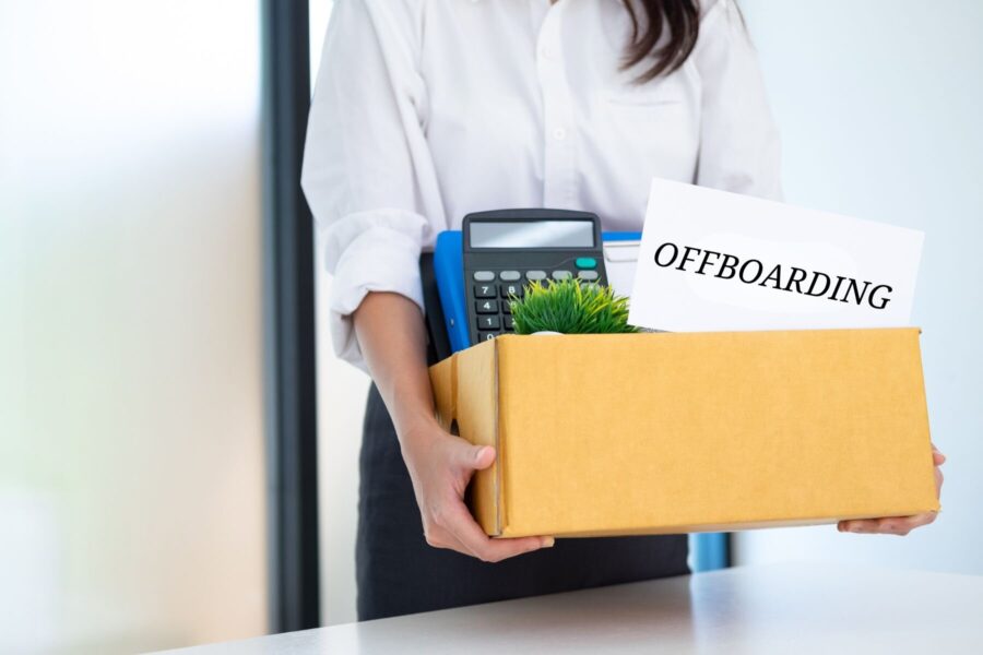 Person hält Karton mit Büromaterial und Schild „OFFBOARDING“.