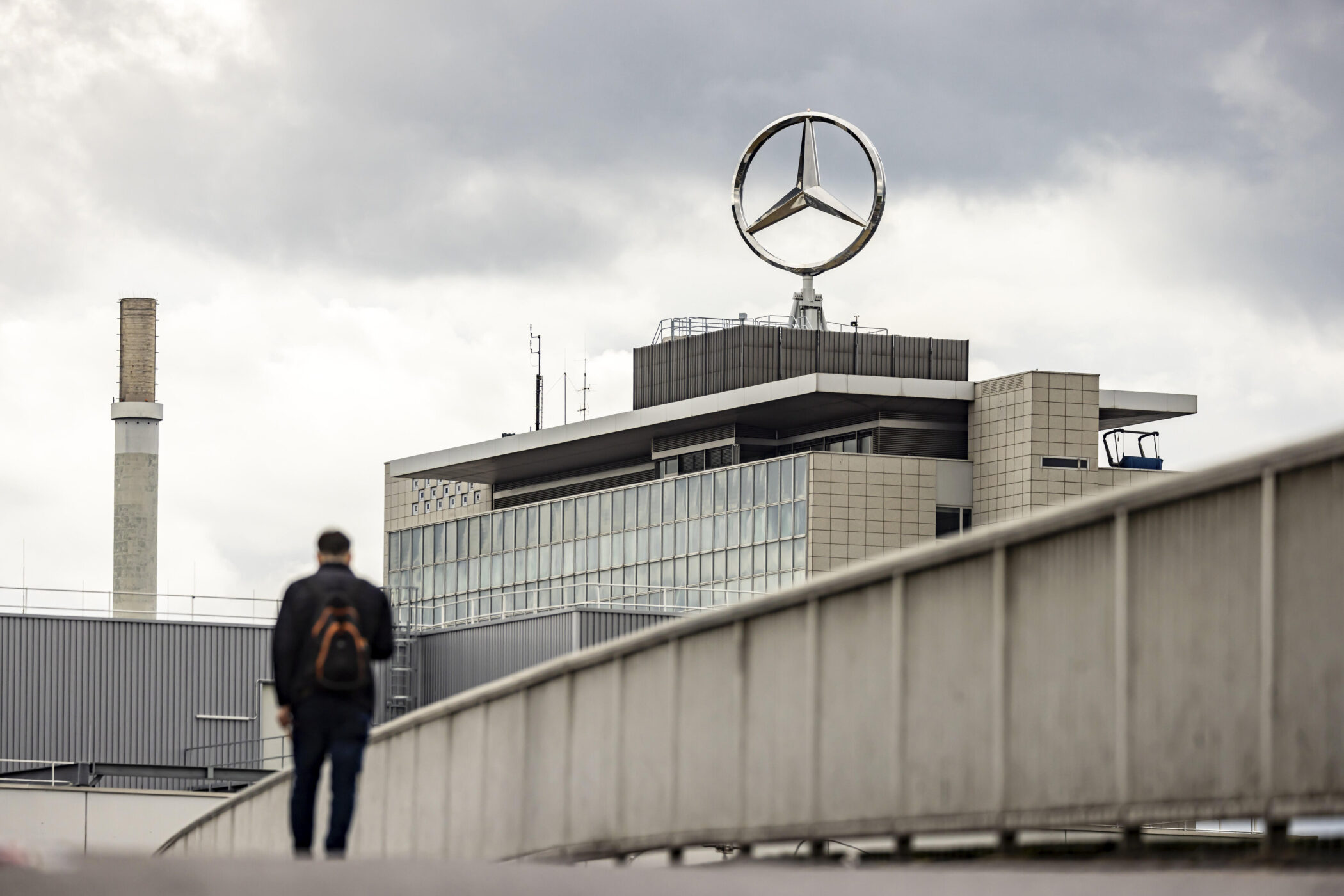 Person mit Rucksack geht auf Brücke, Gebäude mit Mercedes-Stern im Hintergrund.