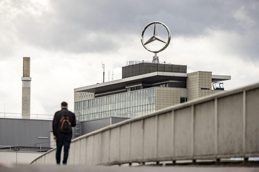 Person mit Rucksack geht auf Brücke, Gebäude mit Mercedes-Stern im Hintergrund.