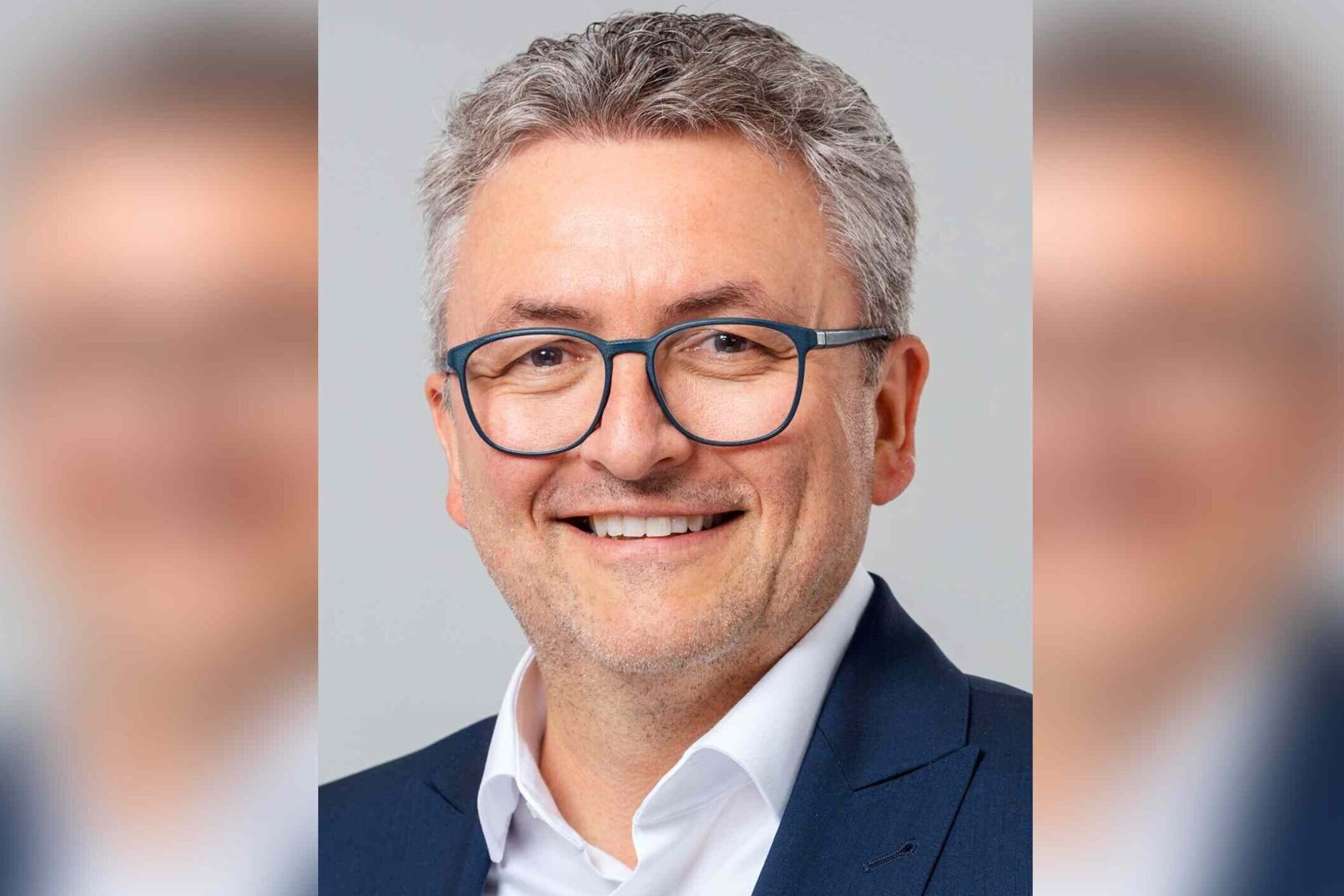 Eine Person mit Brille und grauen Haaren lächelt im Anzug vor neutralem Hintergrund.