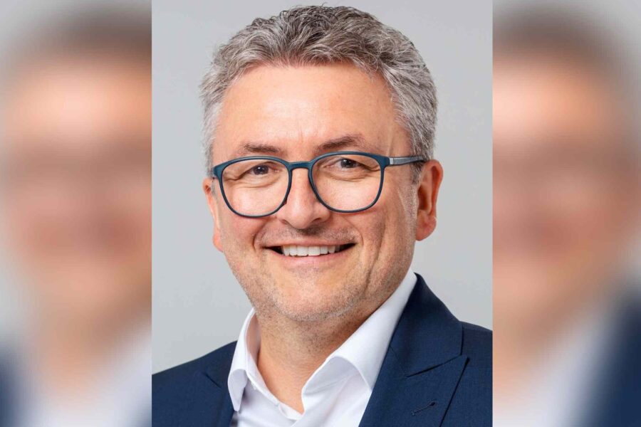 Eine Person mit Brille und grauen Haaren lächelt im Anzug vor neutralem Hintergrund.