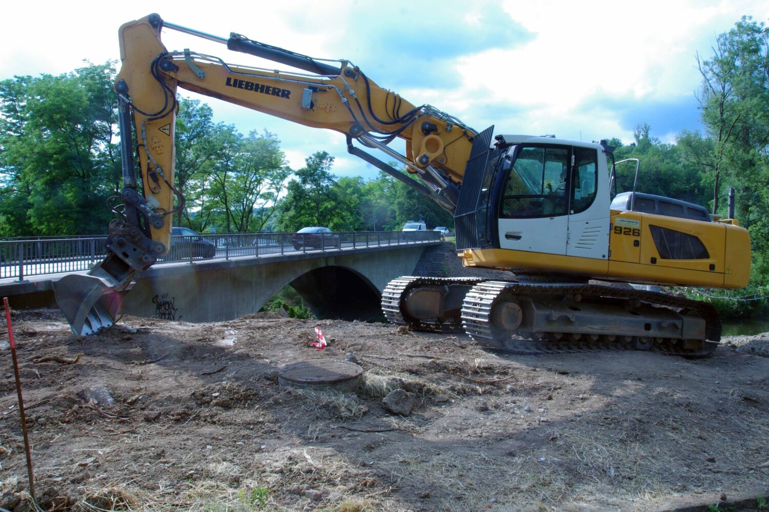 Gelber Bagger auf Baustelle vor Brücke, Bäume im Hintergrund.