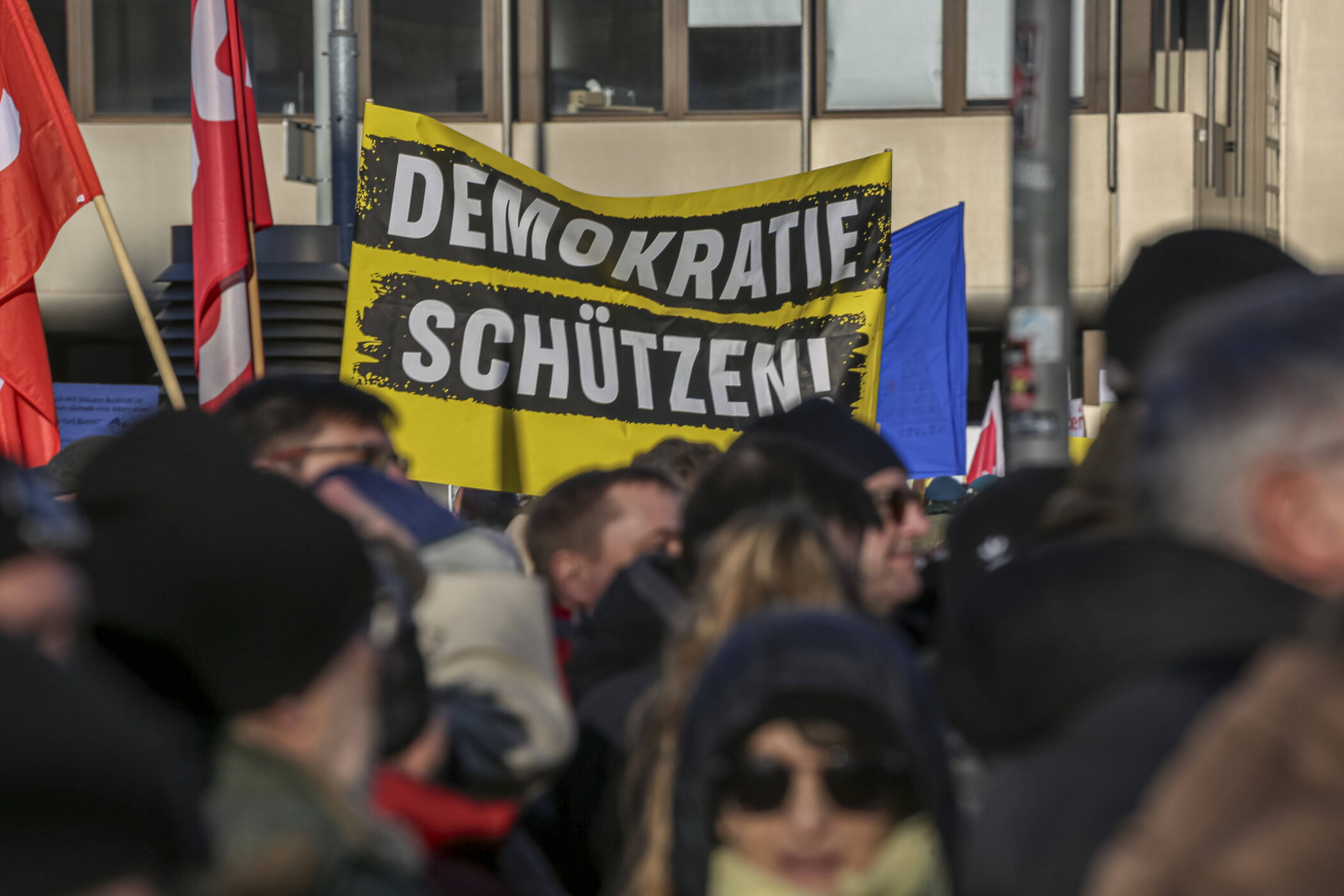 Menschenmenge mit Plakaten, eins sagt "Demokratie schützen", rote Fahnen.