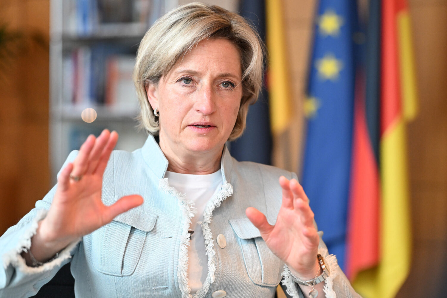 Frau in hellem Blazer gestikuliert vor unscharfen Flaggen im Hintergrund.