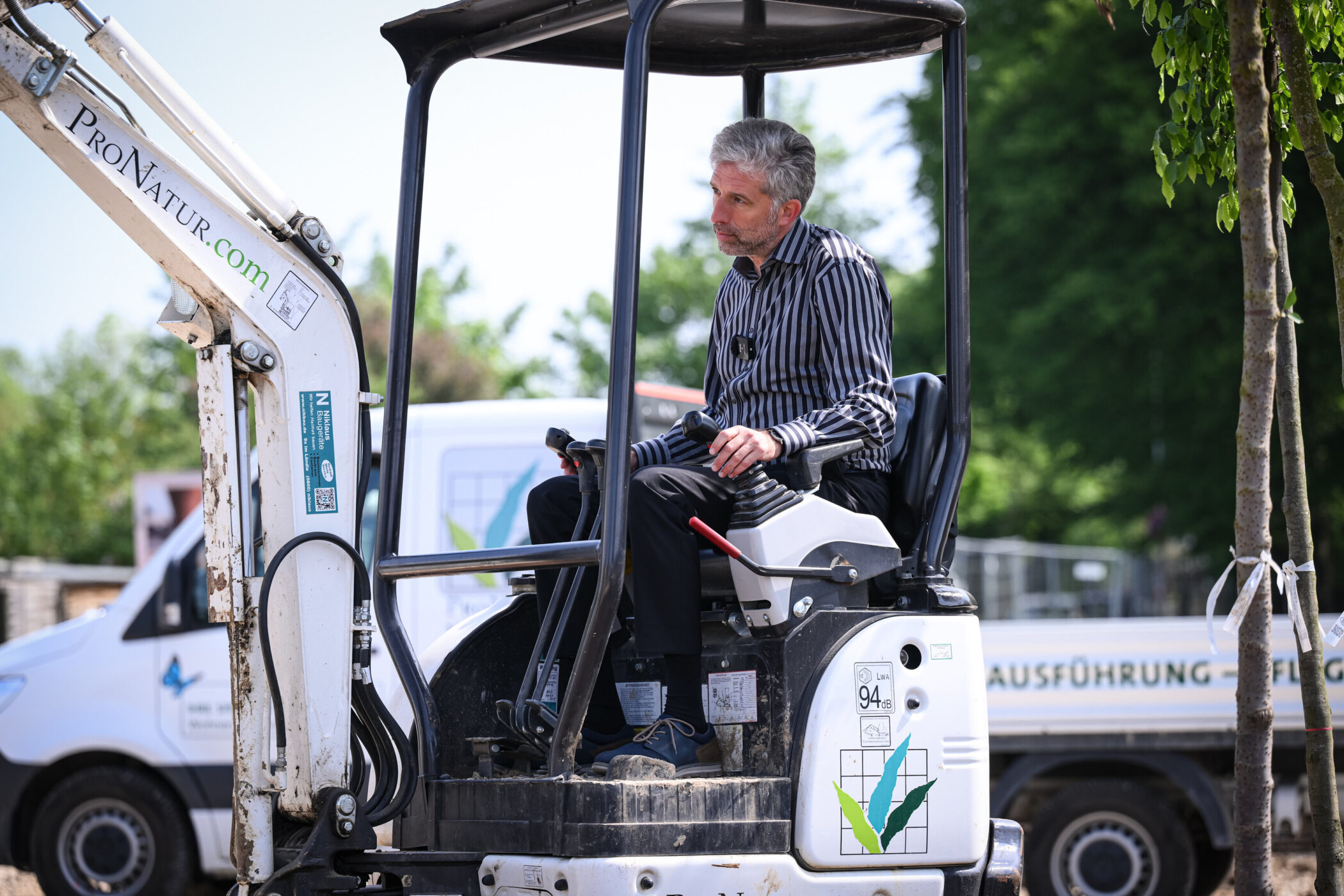 Person in gestreiftem Hemd auf Bagger, im Hintergrund ein Transporter.