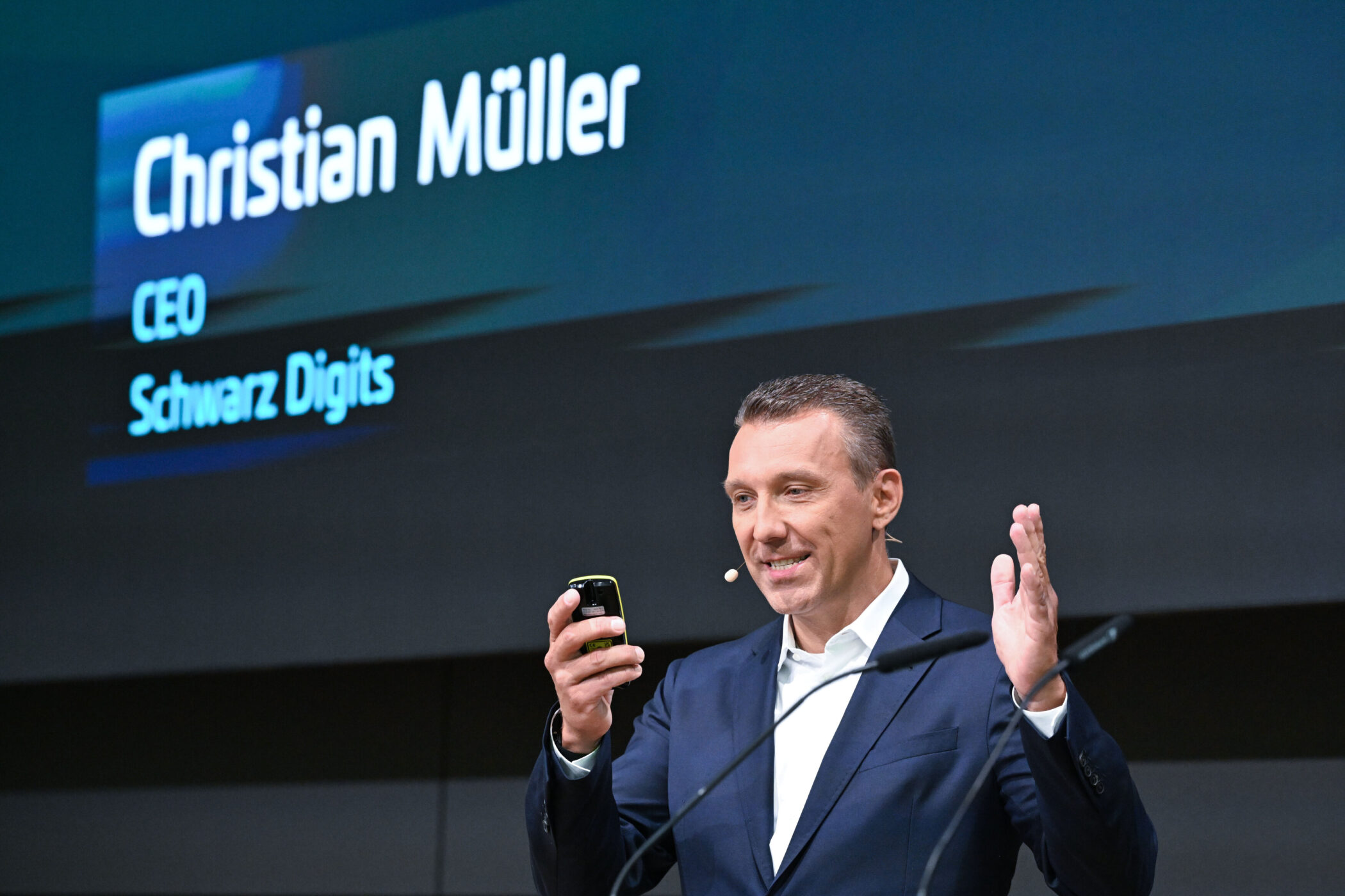 Mann im Anzug mit Mikrofon spricht, hält Handy. Text: "Christian Müller, CEO Schwarz Digits".