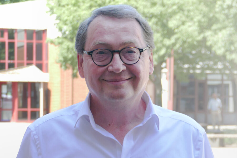 Mann mit Brille und weißem Hemd im Freien, Bäume im Hintergrund.