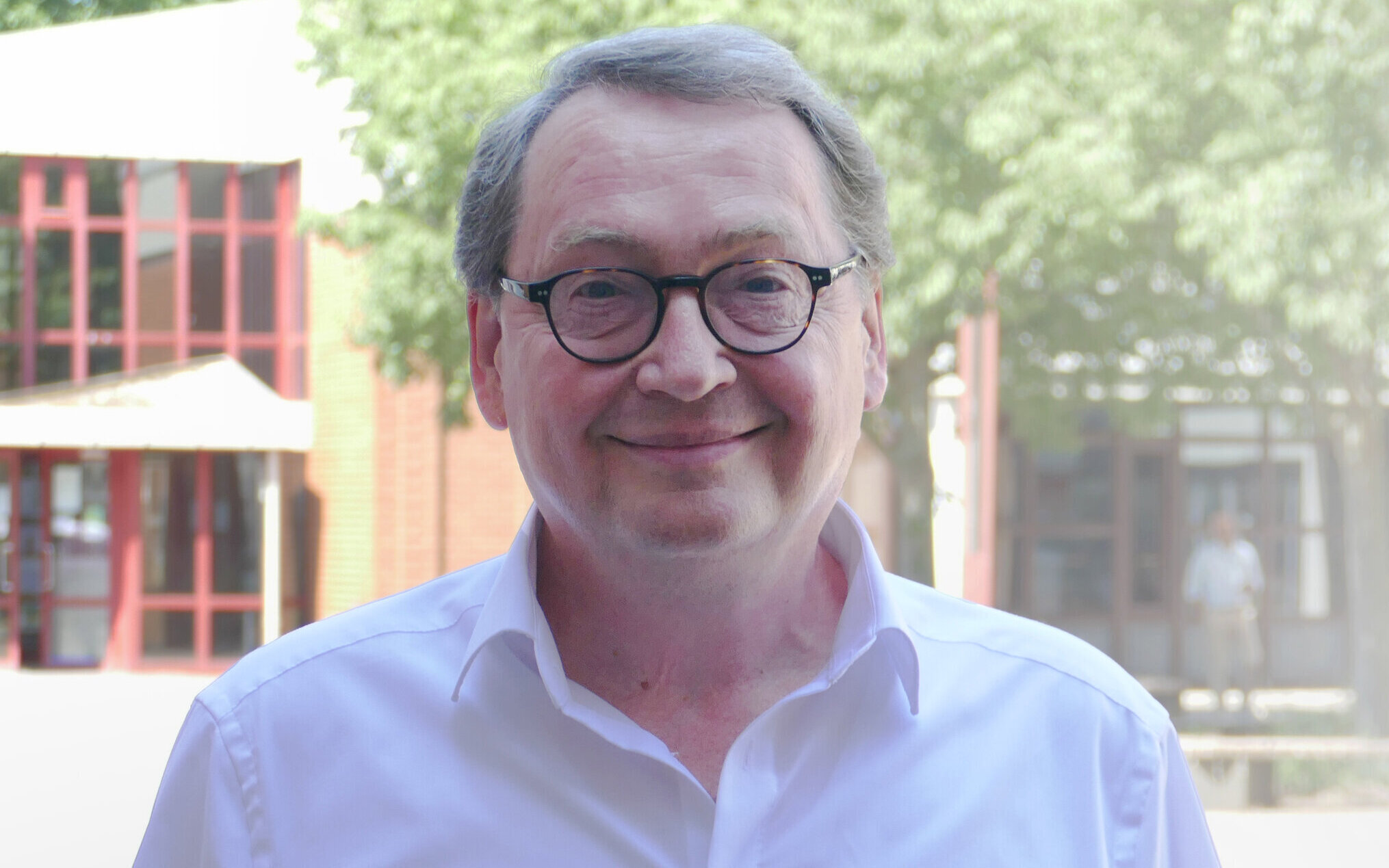 Mann mit Brille und weißem Hemd im Freien, Bäume im Hintergrund.