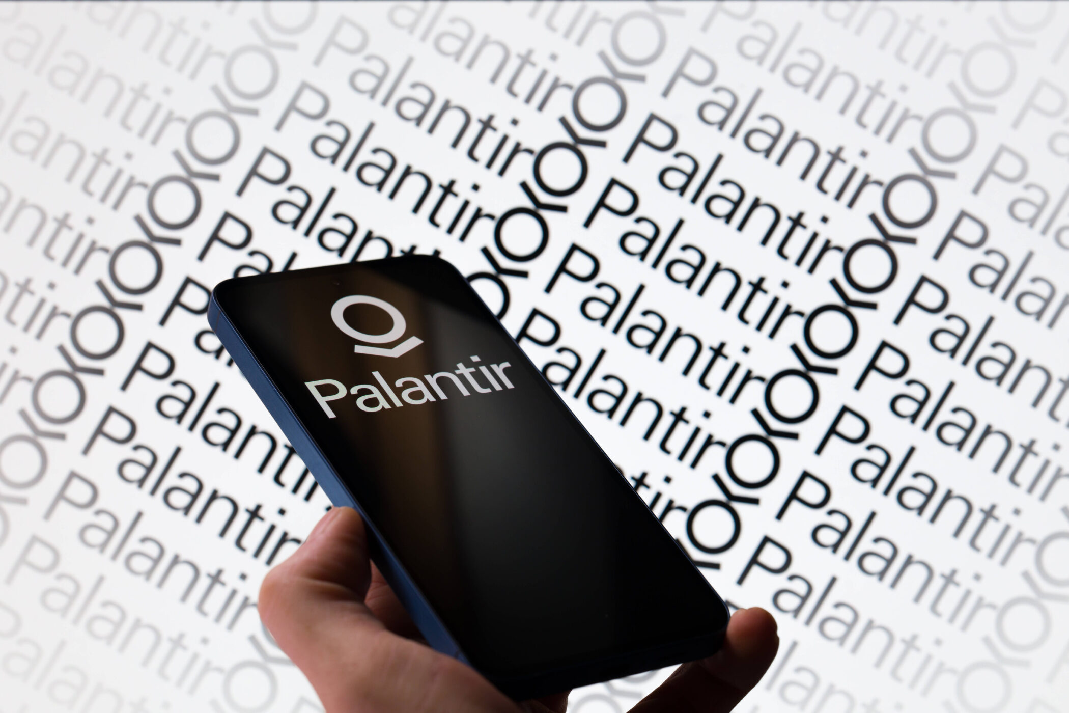 Smartphone mit Palantir-Logo vor Hintergrund mit Palantir-Schriftzug.