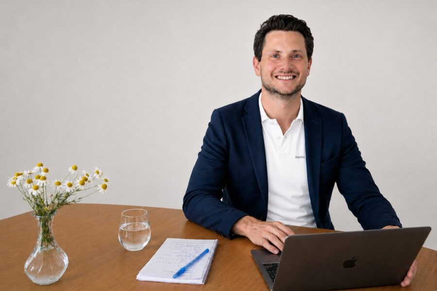 Mann im Anzug am Tisch mit Laptop, Glas, Notizblock und Blumen in Vase.