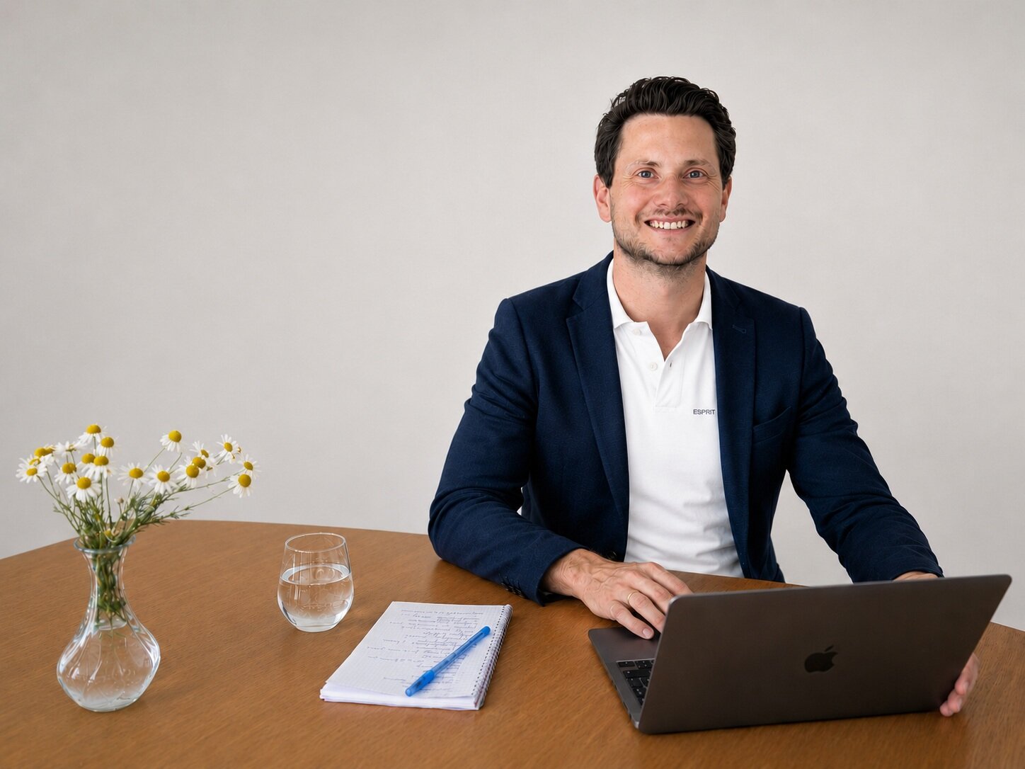 Mann im Anzug am Tisch mit Laptop, Glas, Notizblock und Blumen in Vase.
