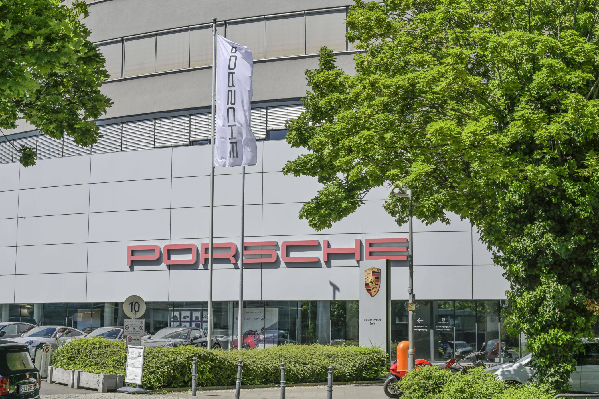 Gebäude mit Porsche-Logo, davor Autos, Flaggenmast, Bushaltestellenschild.