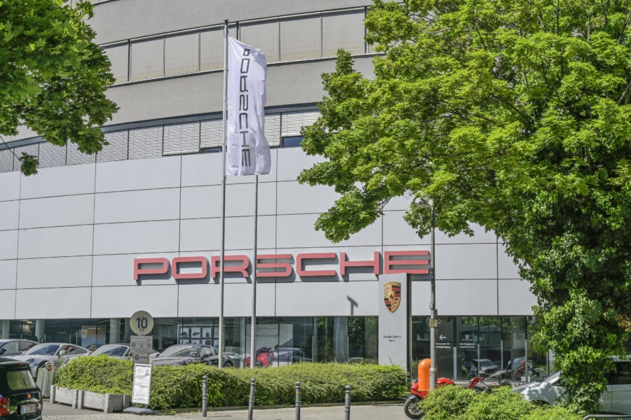 Gebäude mit Porsche-Logo, davor Autos, Flaggenmast, Bushaltestellenschild.