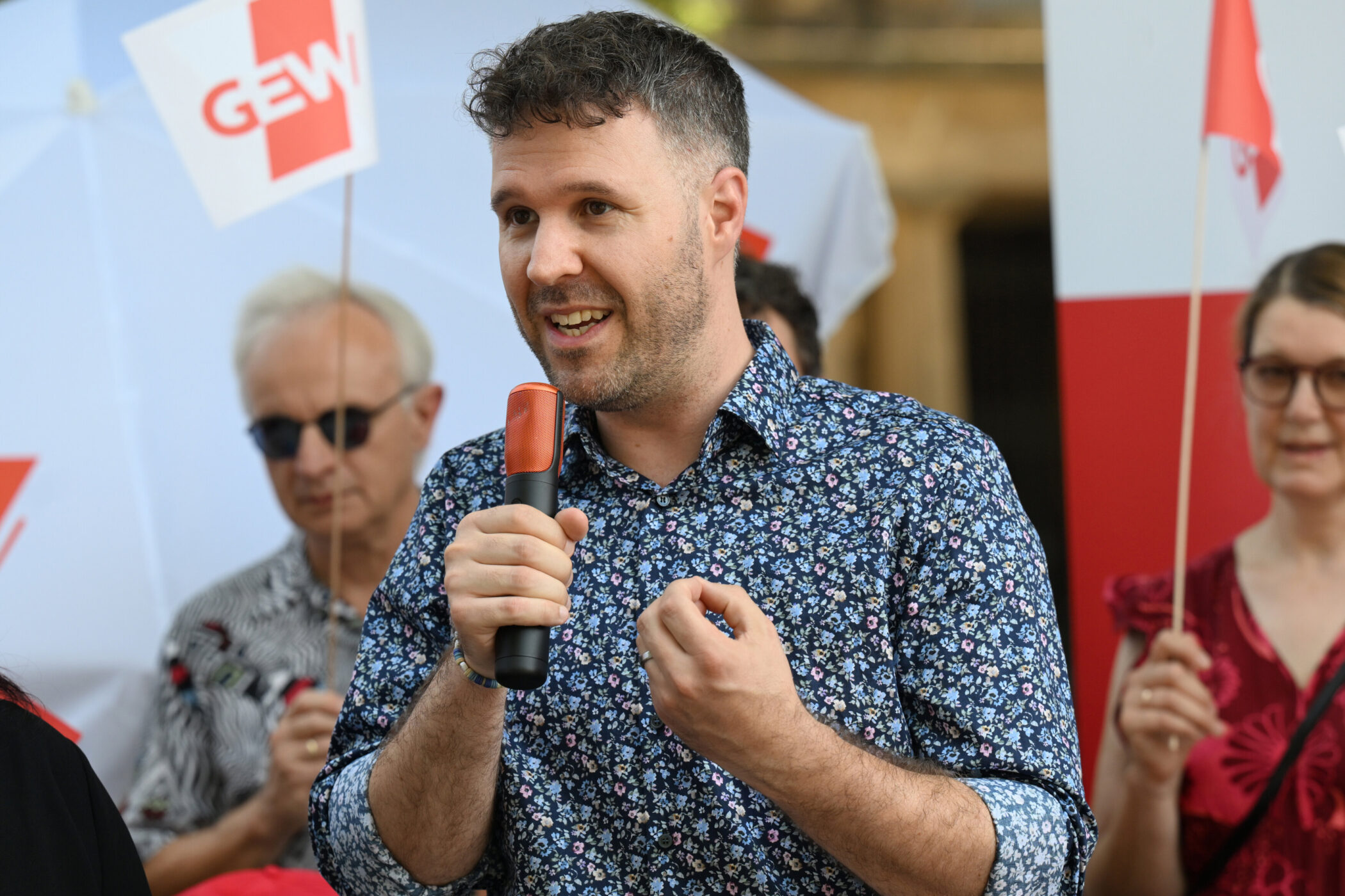 Mann mit Mikrofon, mehrere Personen im Hintergrund mit GEW-Schildern, draußen.