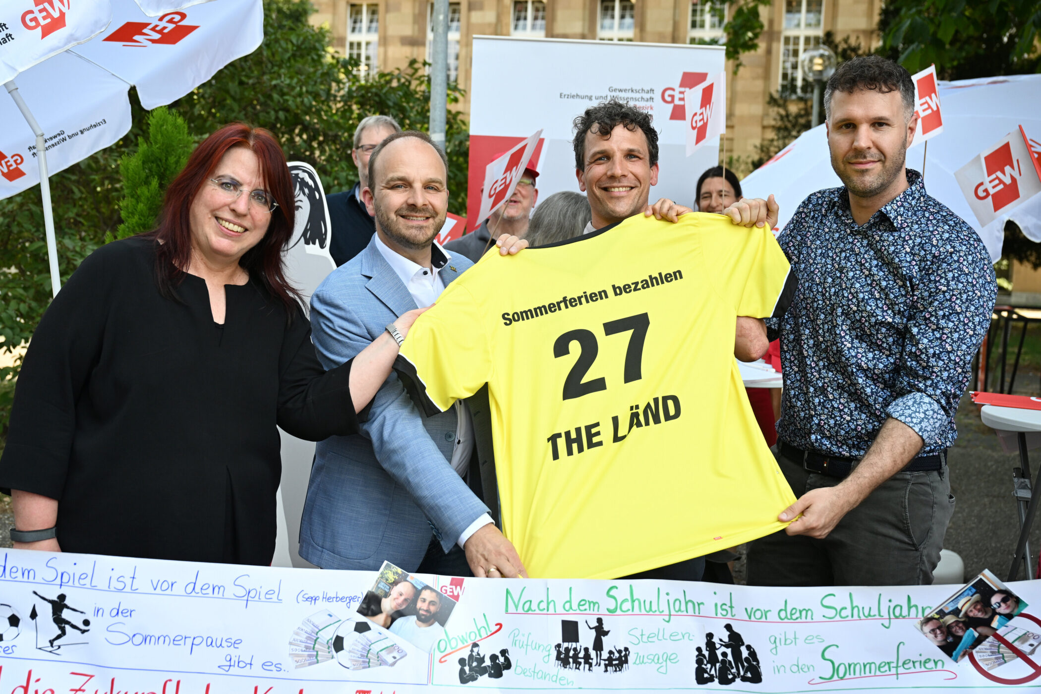 Vier Personen halten ein gelbes Trikot mit der Aufschrift „Sommerferien bezahlen 27 THE LÄND“.