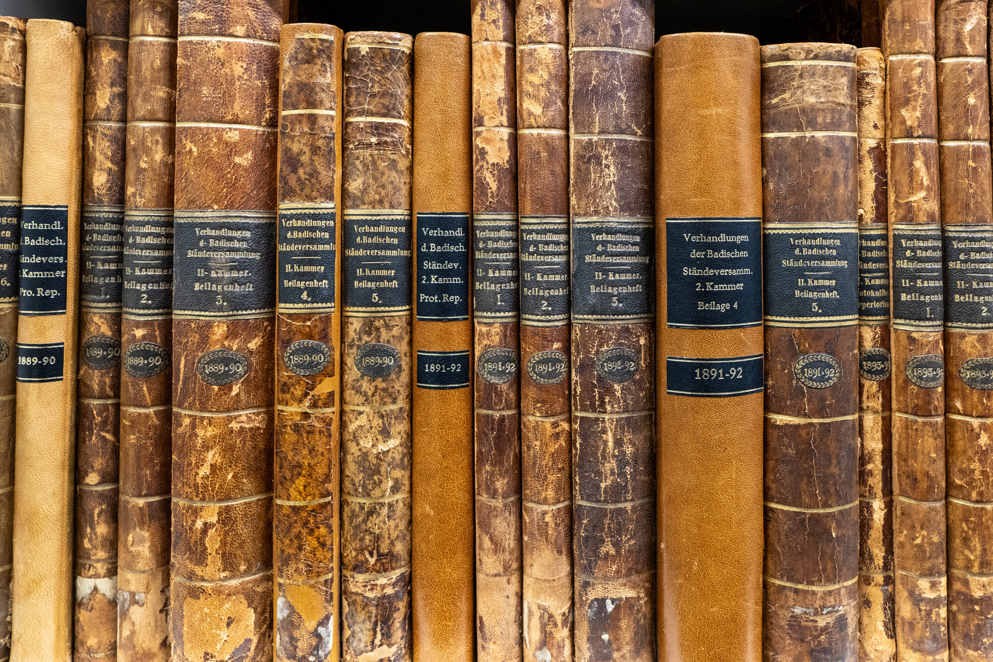 Mehrere alte, braune Bücher mit schwarzen Etiketten auf einem Regal.