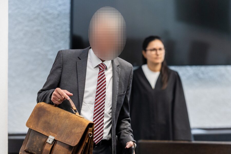 Zwei Personen im Anzug, eine mit Aktentasche, im Hintergrund eine in Robe.
