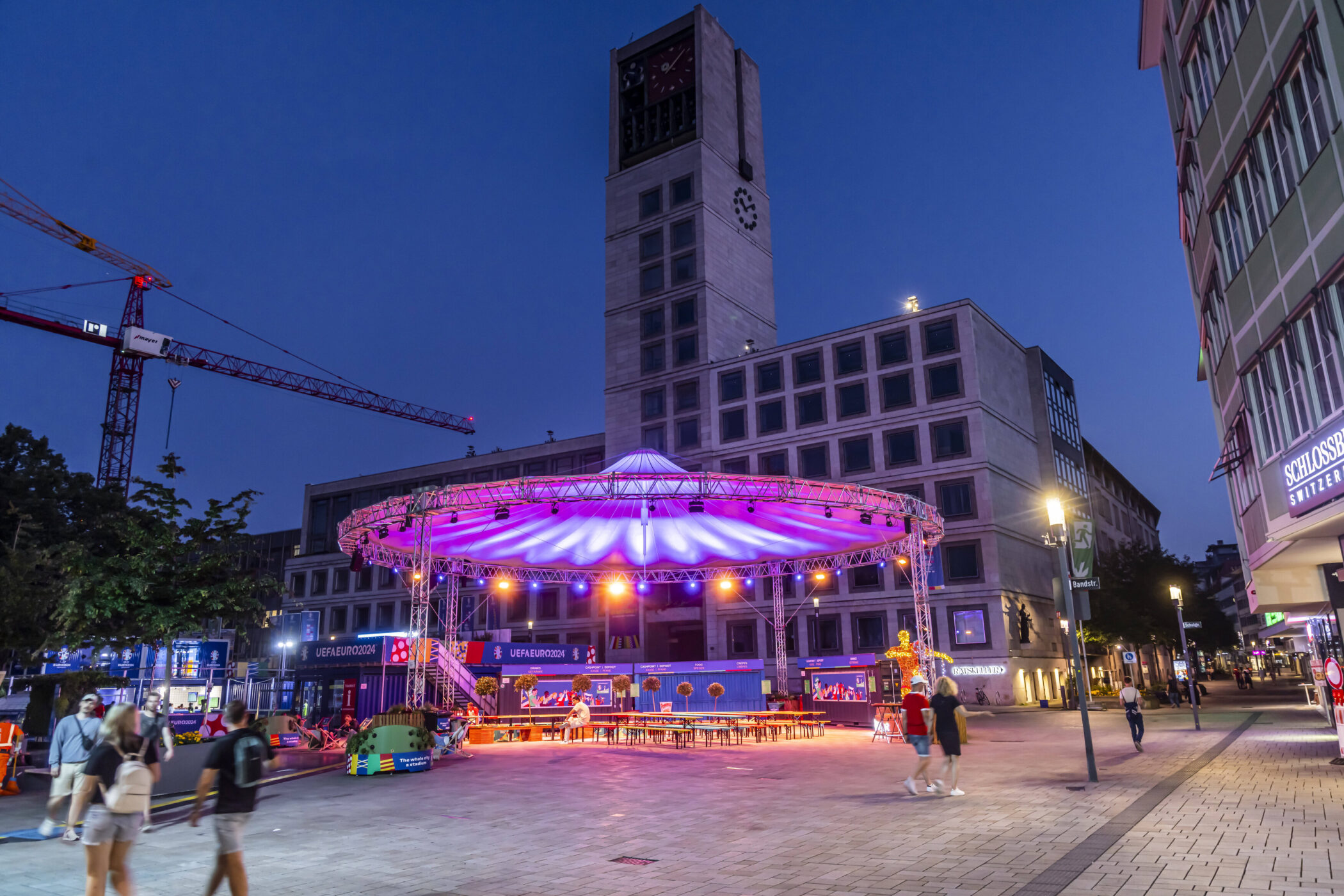 Stadtplatz abends, überdachte Bühne beleuchtet, Menschen spazieren. Uhrturm im Hintergrund.