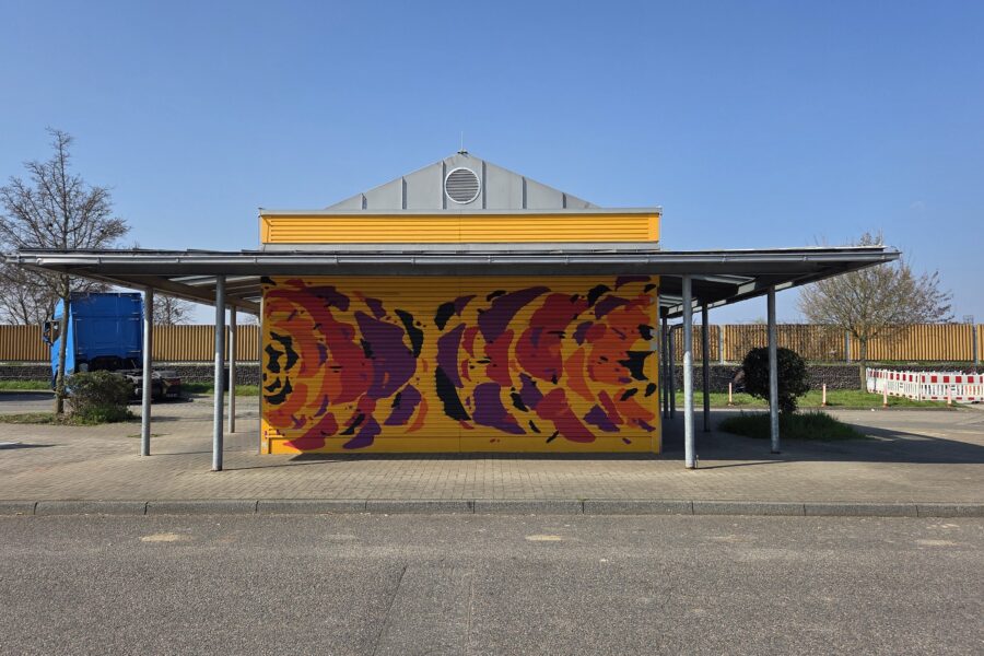 Gelbes Gebäude mit buntem Wandbild auf einer gepflasterten Fläche. Lkw im Hintergrund.
