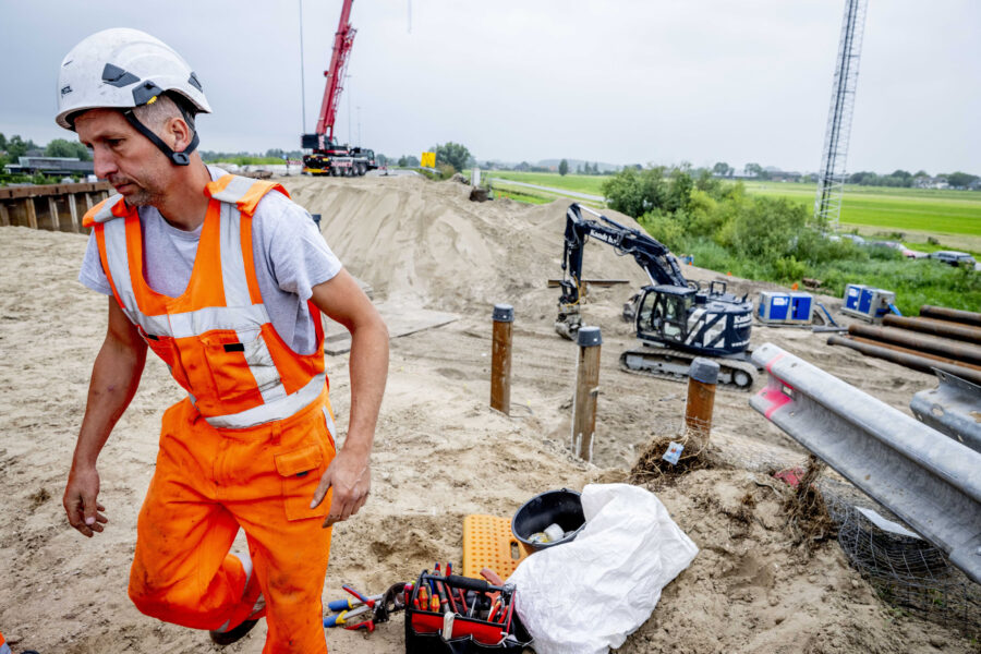 Arbeiter in oranger Kleidung auf Baustelle mit Bagger, Werkzeugkiste und Sandhaufen.