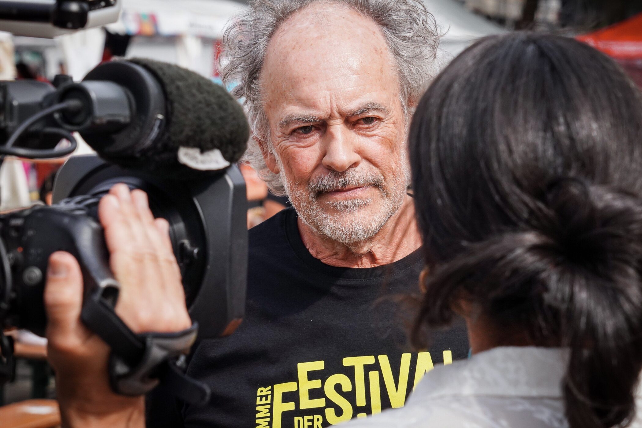 Zwei Personen, Kamera, Mann mit grauem Haar und Bart, T-Shirt-Text: "FESTIVAL".