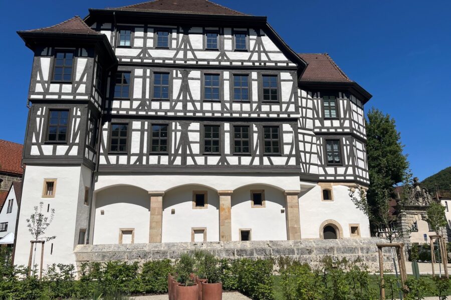 Fachwerkgebäude mit weißer Fassade, dahinter blauer Himmel, vorne Gartenanlage.