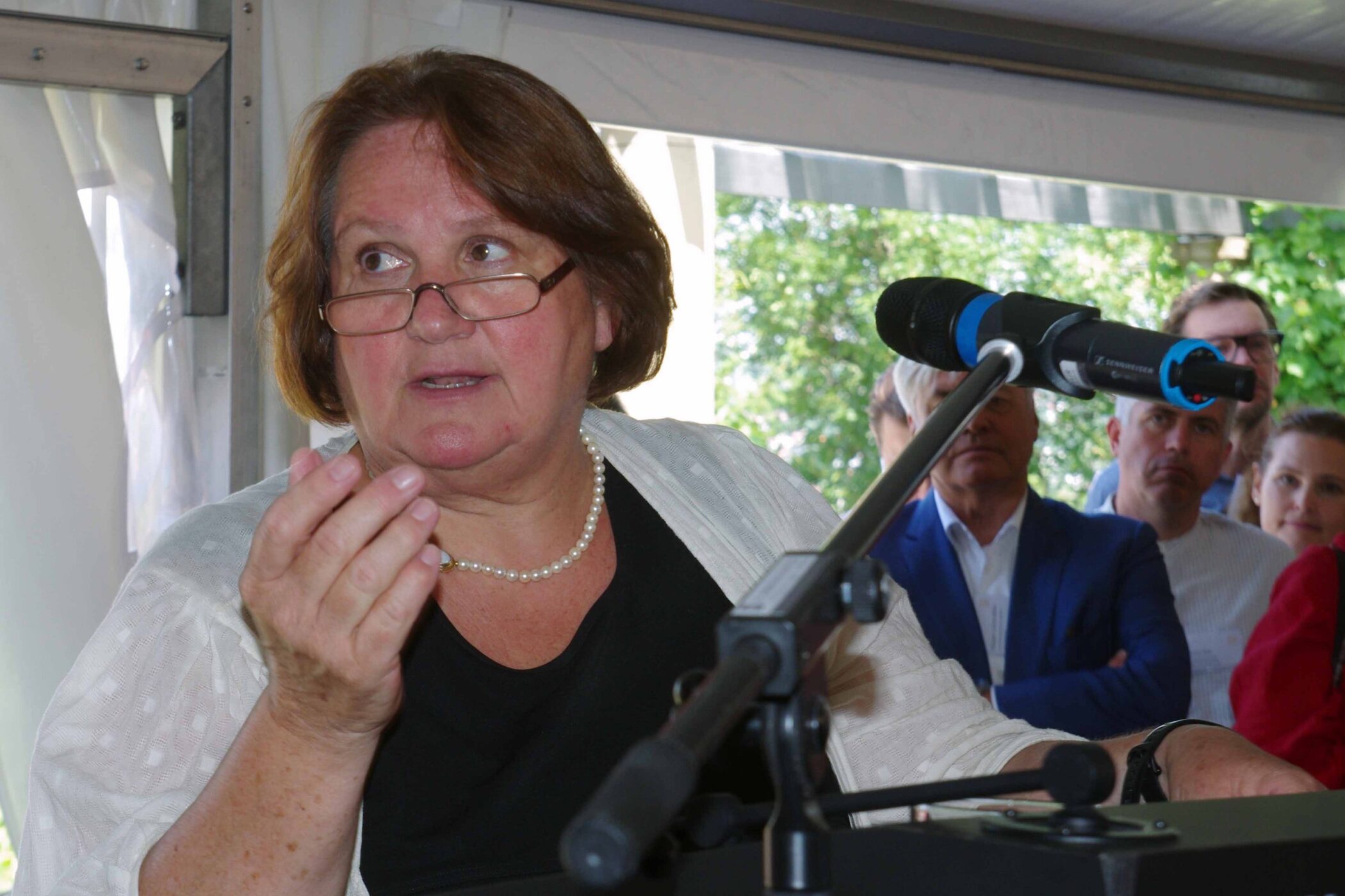 Frau am Mikrofon, Publikum im Hintergrund, Lichtdurchflutete Umgebung.