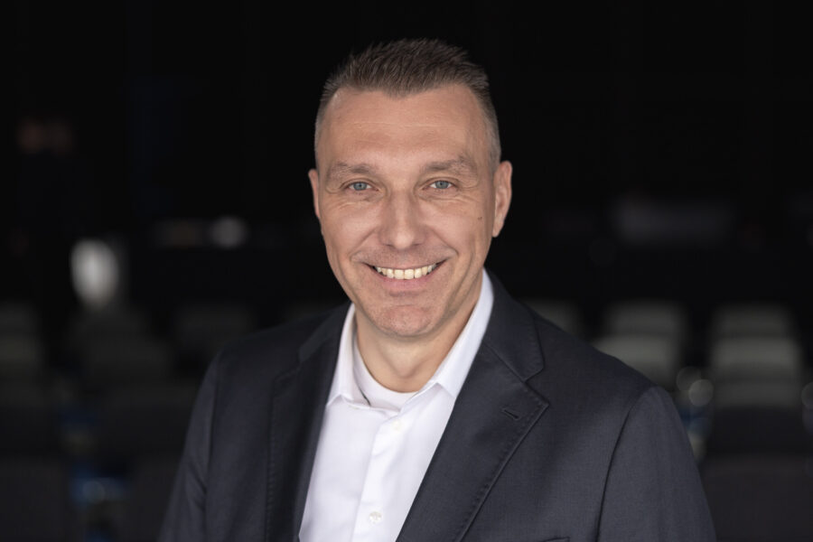 Mann im Anzug lächelt vor unscharfem Hintergrund.