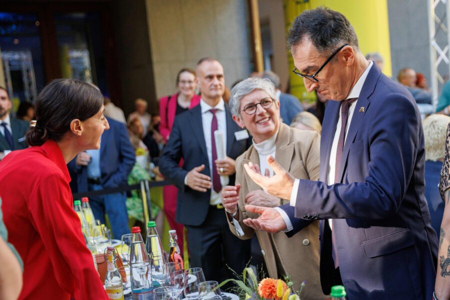 Mehrere Personen in Anzügen und Jacken, sprechen an einem Tisch mit Getränken.