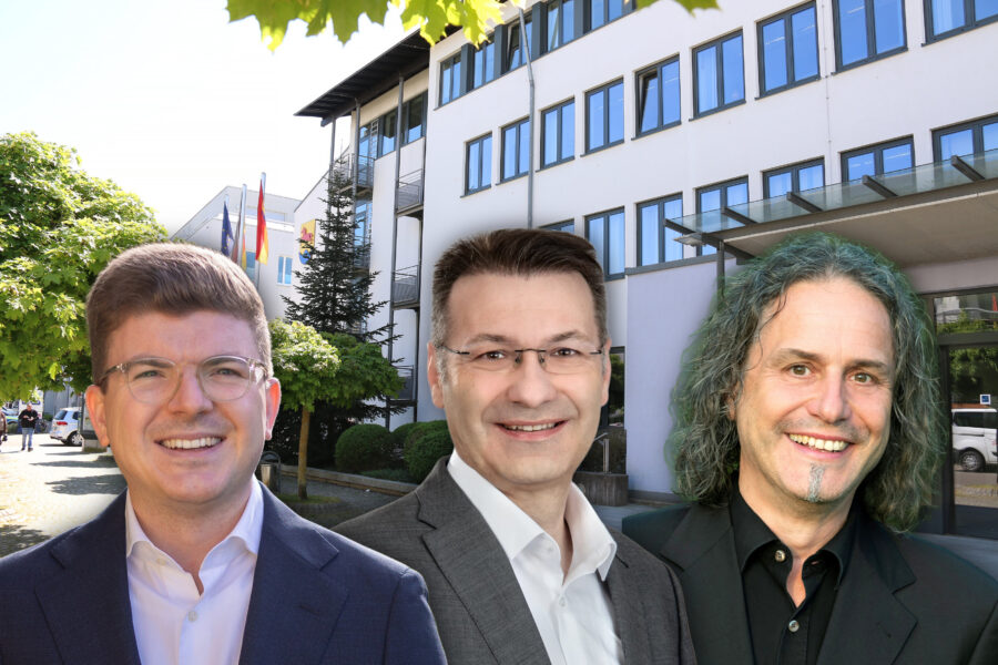 Drei Männer lächeln, im Hintergrund ein Gebäude mit Fenstern und Bäumen.
