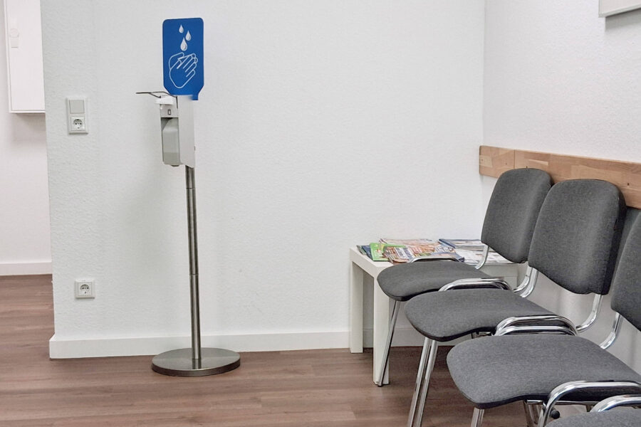 Desinfektionsmittelständer mit Schild, vier graue Stühle, Tisch mit Zeitschriften.