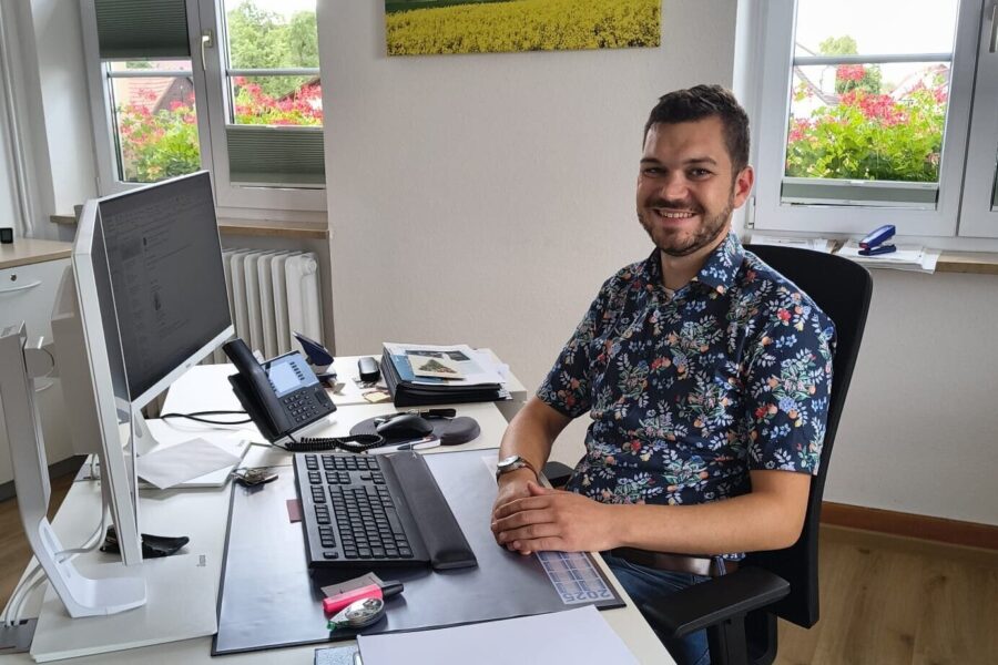 Mann am Schreibtisch mit Computer und Telefon, Fenster im Hintergrund.