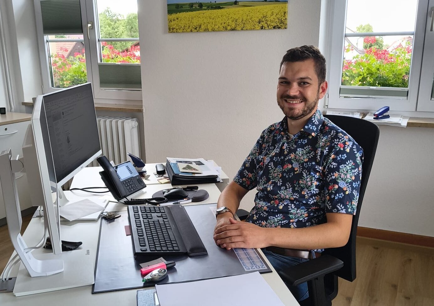 Mann am Schreibtisch mit Computer und Telefon, Fenster im Hintergrund.