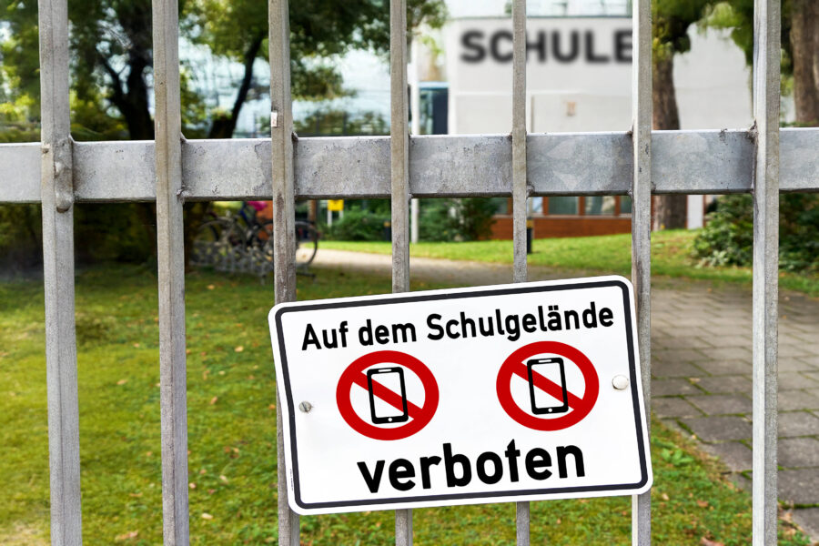 Metallzaun mit Schild: "Auf dem Schulgelände verboten", zeigt durchgestrichene Handys.