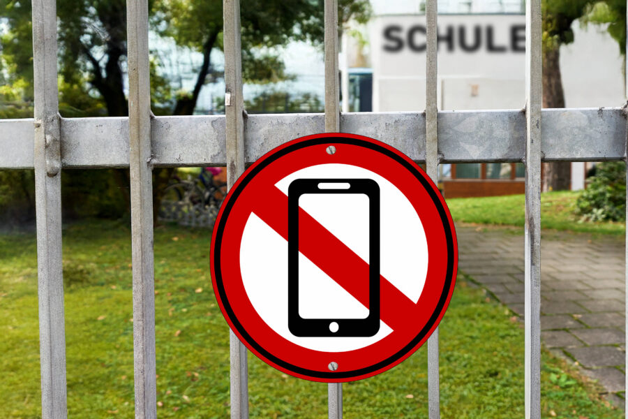 Verbotszeichen für Handys an einem Metallzaun, Gebäude mit "SCHULE" im Hintergrund.