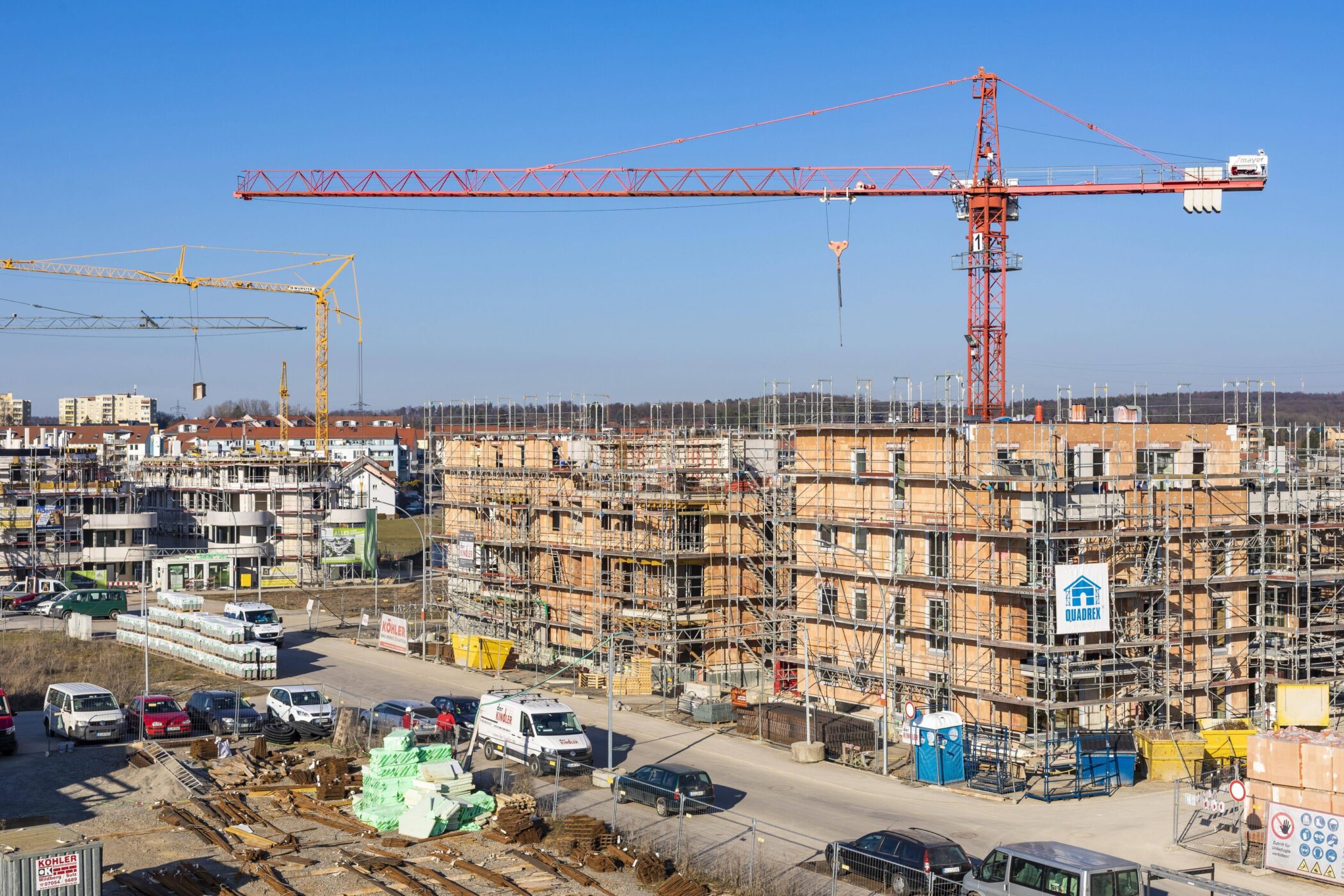 Baustelle mit Baukränen, Gerüsten und parkenden Autos unter blauem Himmel.