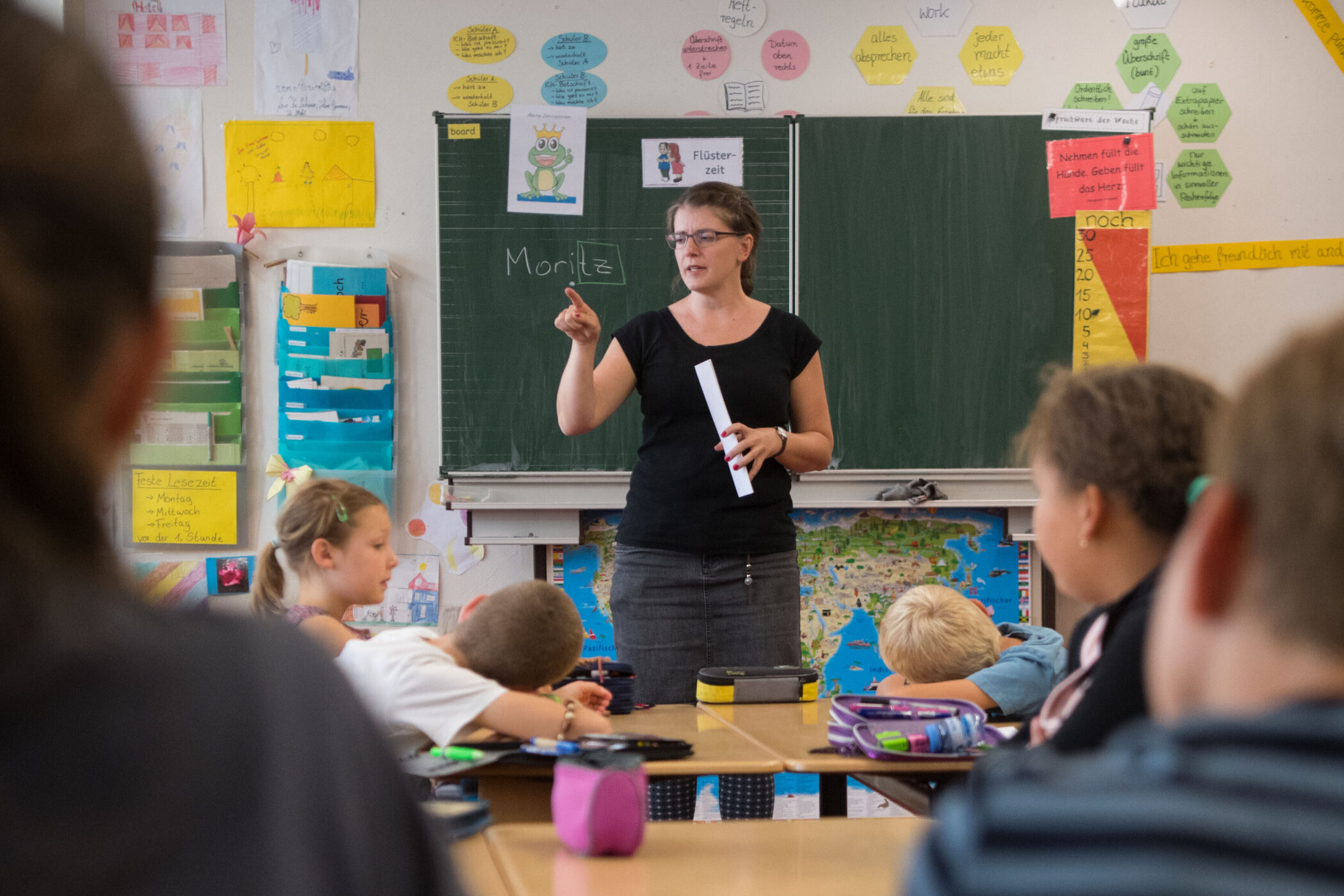 Lehrerin steht vor Tafel, Kinder an Tischen, Tafel mit "Moritz" beschrieben.