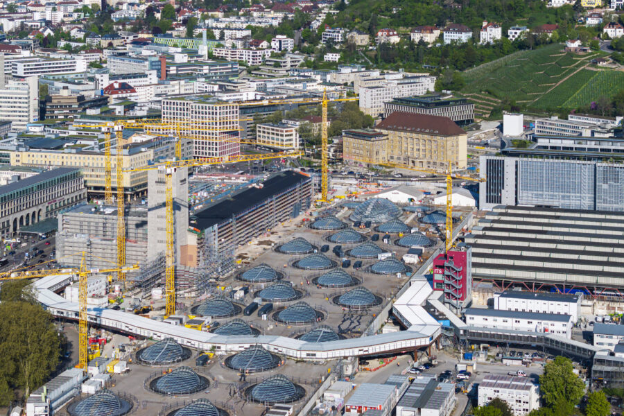 Stadtansicht mit großen Baustellen, Kränen und Gebäuden mit Glasdächern, grüner Umgebung.