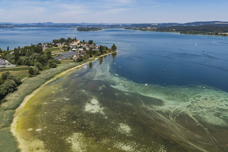 See mit Insel und Dorf, umgeben von Bäumen, klarer Himmel, einige Boote im Wasser.