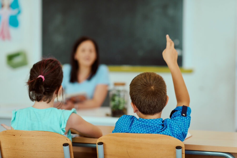 Zwei Kinder sitzen an Tischen, eines hebt die Hand, Lehrerin im Hintergrund.