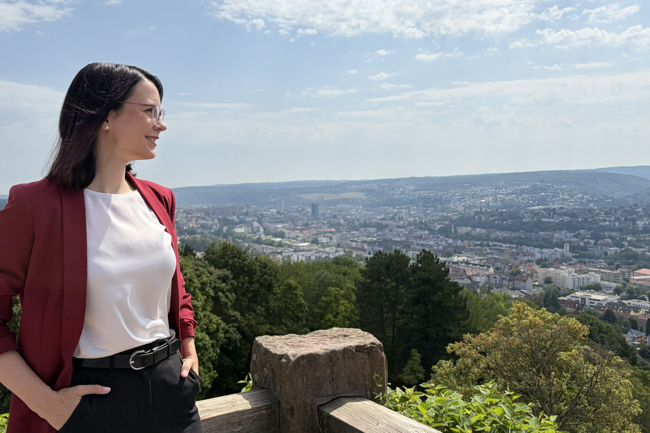 Eine Person steht vor einem Stadtpanorama, trägt Blazer und blickt zur Seite.