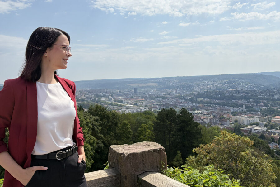Eine Person steht vor einem Stadtpanorama, trägt Blazer und blickt zur Seite.