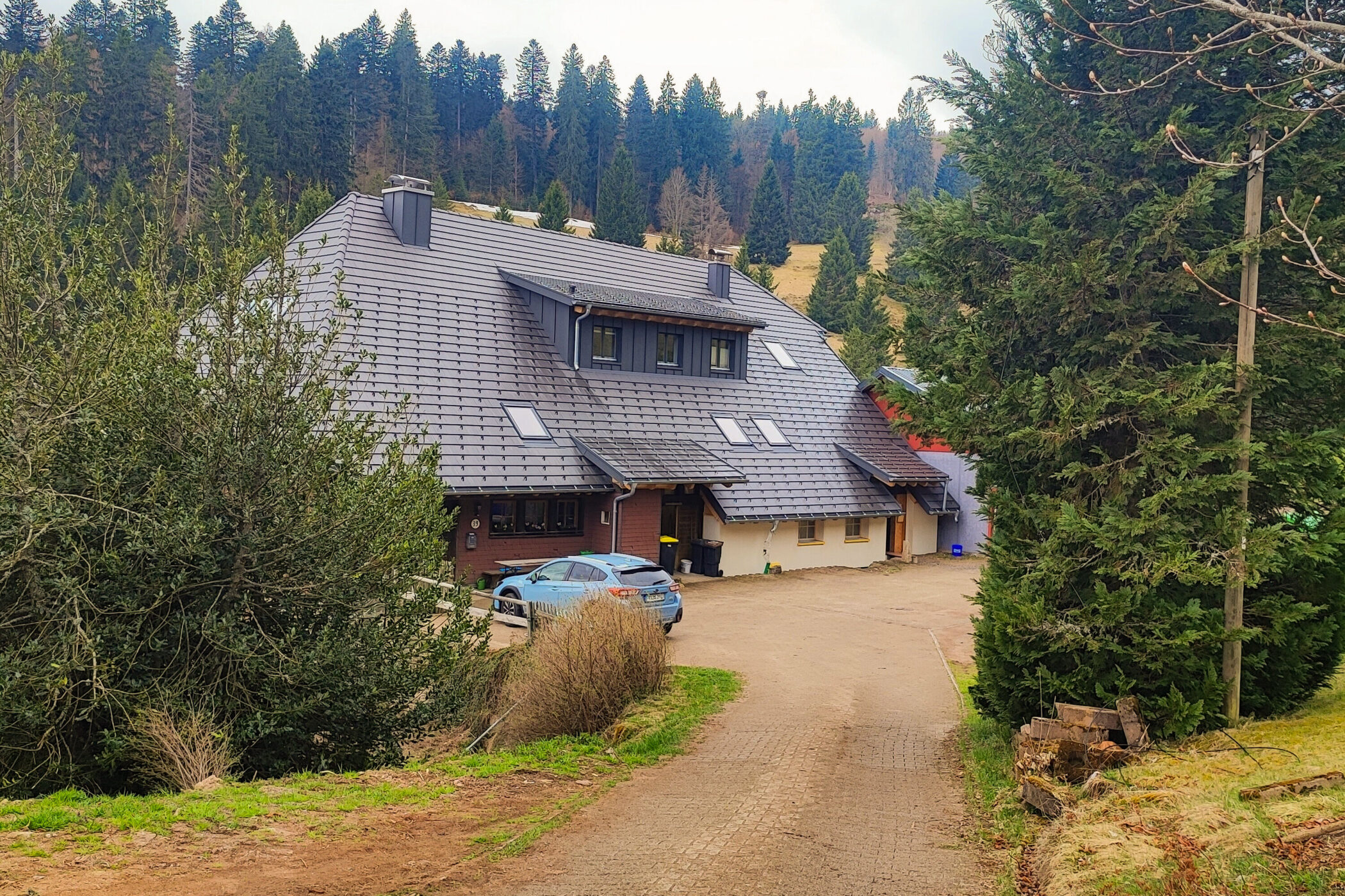Großes Haus mit schrägem Dach im Wald, zwei Autos, Kiesweg, umgeben von Bäumen.