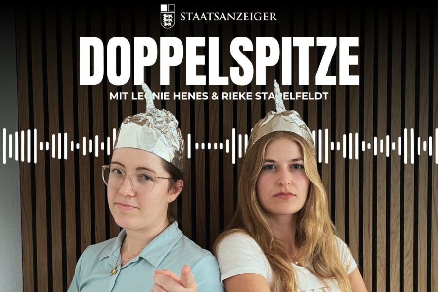 Zwei Personen mit Aluhüten vor Holzpaneelen. Text: STAATSANZEIGER DOPPELSPITZE.