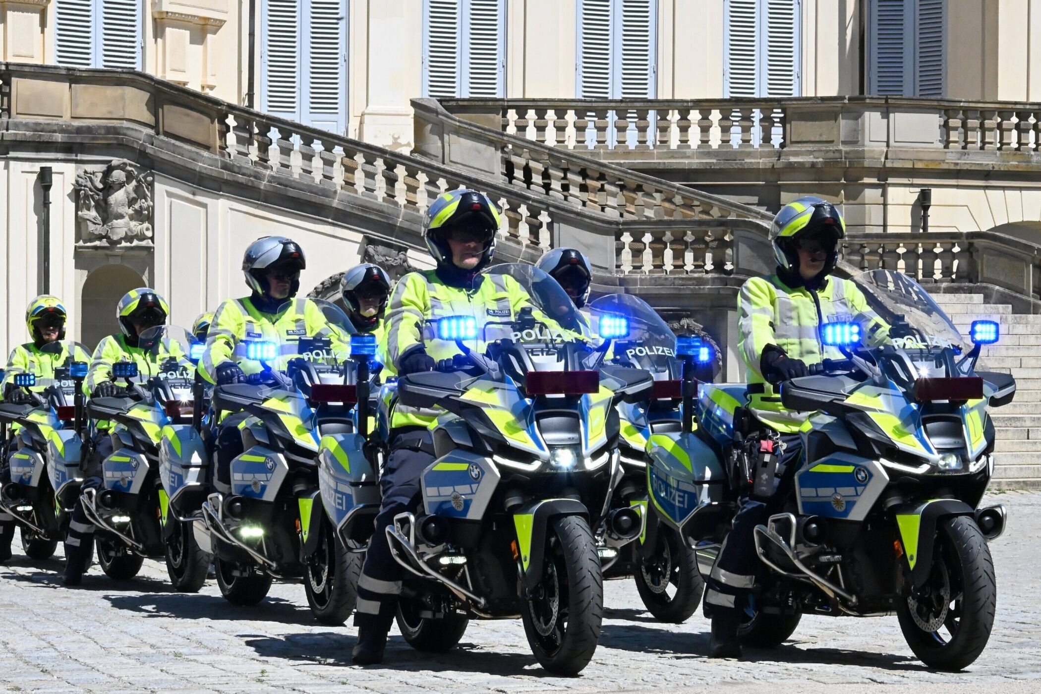 Sechs Polizisten fahren auf Motorrädern in gelben Jacken vor historischem Gebäude.
