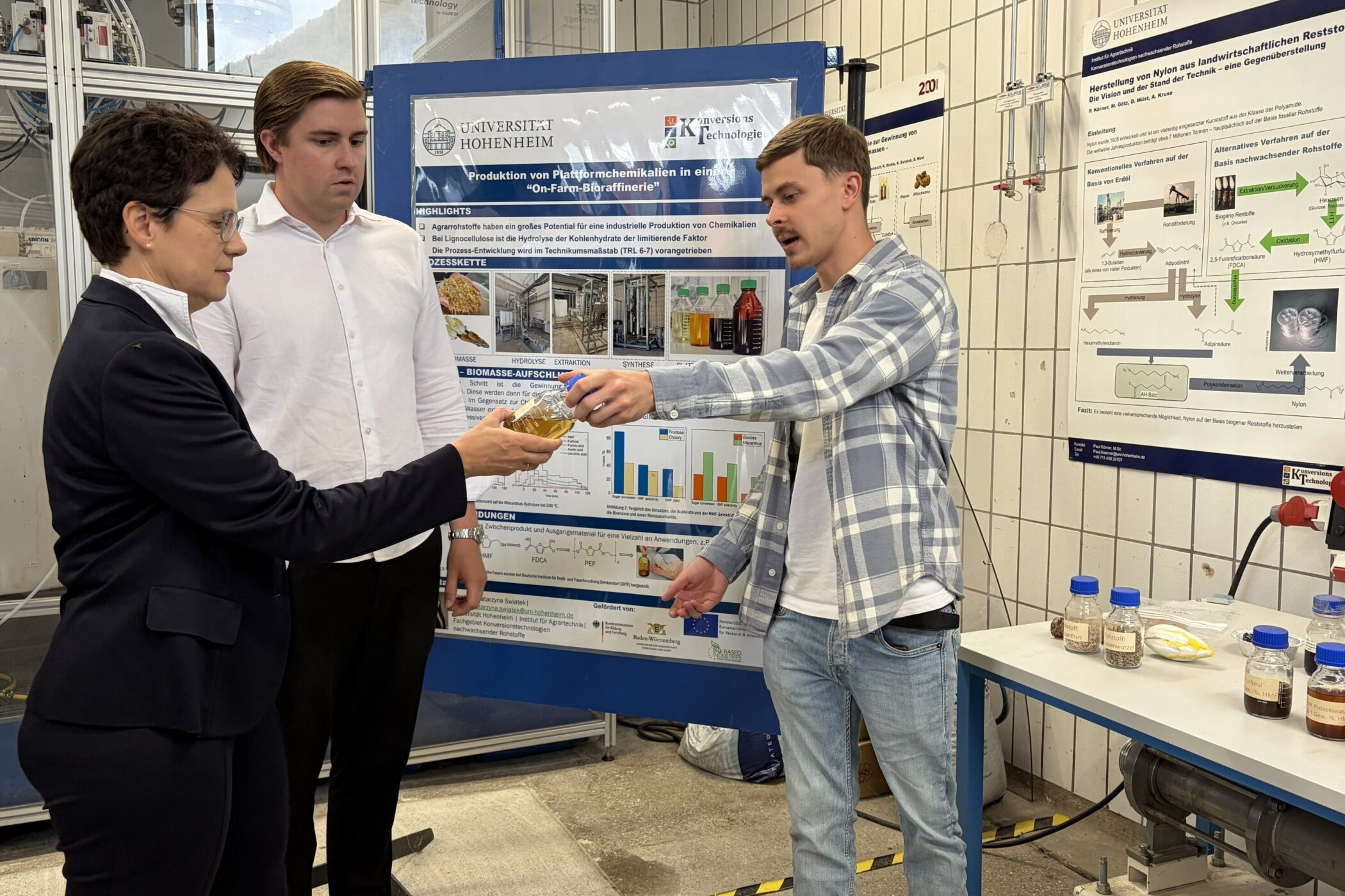 Drei Personen, eine gibt einem anderen eine Flasche, wissenschaftliche Poster im Hintergrund.